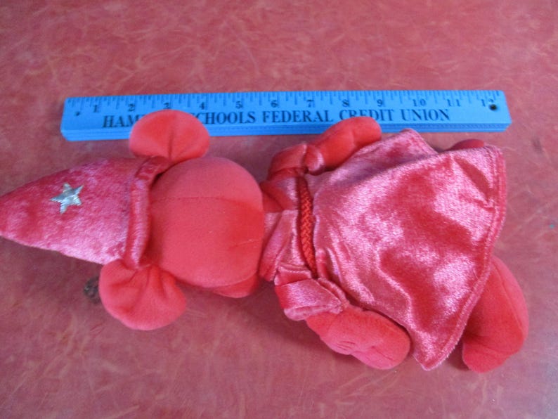 May include: A red plush toy, possibly a costume, with a pointed hat featuring a silver star and a cape. A blue ruler with white text is in the background. The text on the ruler reads "HAM SCHOOLS FEDERAL CREDIT UNION".