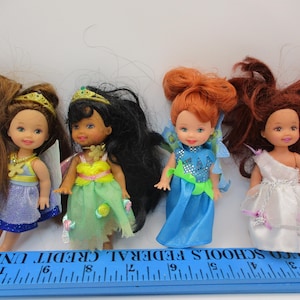 May include: Four small fairy dolls with long hair and colorful outfits. The dolls have wings and tiaras. One doll wears a blue dress, another a green dress, a third a blue dress, and the last a white dress. A blue ruler is at the bottom.