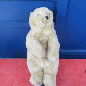 Puede incluir: Un peluche de oso polar con pelaje color crema. El oso está de pie con los brazos a los lados. Tiene una nariz negra y ojos pequeños y oscuros. El juguete está sobre una superficie roja con un fondo azul.