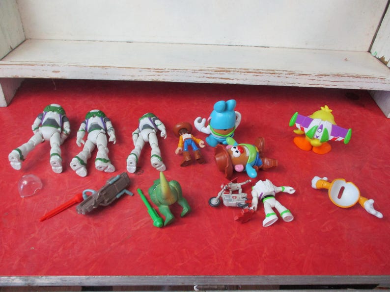 May include: A selection of Toy Story character figures, including Buzz Lightyear, Woody, Mr. Potato Head, and others. The toys are displayed on a red surface, with a white shelf in the background. Various accessories are also present.