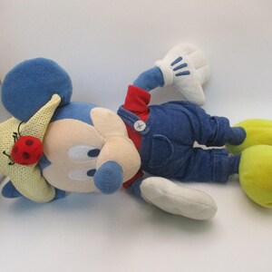 May include: A plush Mickey Mouse doll wearing blue denim overalls, a red shirt, and yellow shoes. The doll has a blue head and ears, white gloves, and a straw hat with a red ladybug. The doll is lying down.
