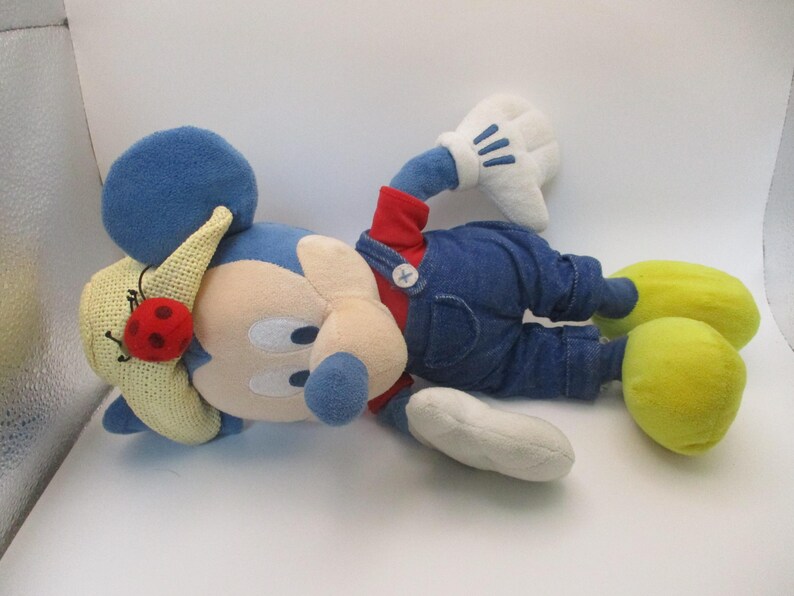 May include: A plush Mickey Mouse doll wearing blue denim overalls, a red shirt, and yellow shoes. The doll has a blue head and ears, white gloves, and a straw hat with a red ladybug. The doll is lying down.