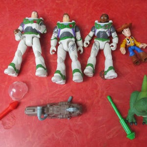 May include: A selection of Toy Story character figures and accessories. The image shows three Buzz Lightyear figures in white and green space suits, Woody, a green dinosaur, and a red rocket. The toys are arranged on a red surface.