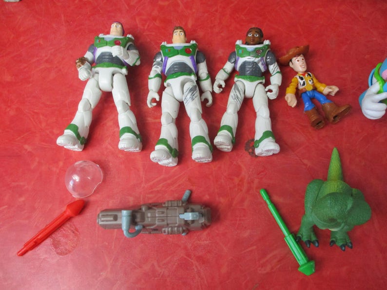 May include: A selection of Toy Story character figures and accessories. The image shows three Buzz Lightyear figures in white and green space suits, Woody, a green dinosaur, and a red rocket. The toys are arranged on a red surface.