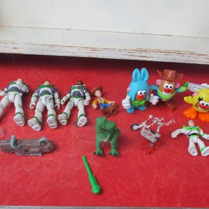 May include: A selection of Toy Story character figurines, such as Buzz Lightyear, Woody, and Mr. Potato Head, displayed on a red surface. The collection includes a green dinosaur and other accessories, creating a playful arrangement.