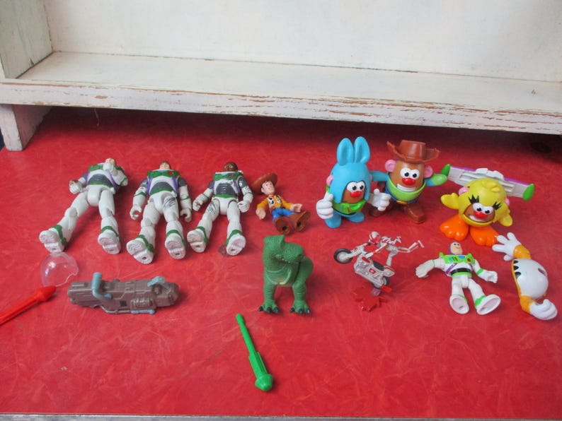May include: A selection of Toy Story character figurines, such as Buzz Lightyear, Woody, and Mr. Potato Head, displayed on a red surface. The collection includes a green dinosaur and other accessories, creating a playful arrangement.