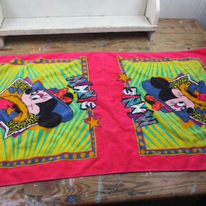 May include: A pink rectangular towel featuring two identical cartoon character designs. Each design includes a cartoon mouse in a blue hat and yellow jacket, set against a yellow and green background. The word "MINNIE" is also displayed.