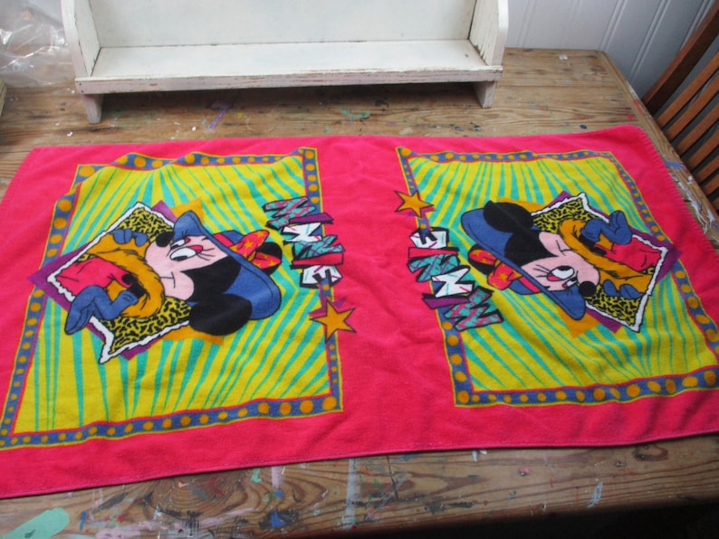 May include: A pink rectangular towel featuring two identical cartoon character designs. Each design includes a cartoon mouse in a blue hat and yellow jacket, set against a yellow and green background. The word "MINNIE" is also displayed.