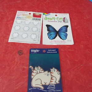 May include: Three iron-on fabric appliques on a red surface. The appliques include white flower shapes, a blue butterfly, and three cats. The butterfly applique is labeled "Delta Iron-On Transfer."