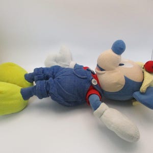 May include: A plush toy of a cartoon character, lying on its back. The character has a blue head and hat, a red nose, and a yellow and red hat. It wears blue overalls, a red shirt, white gloves, and yellow shoes.