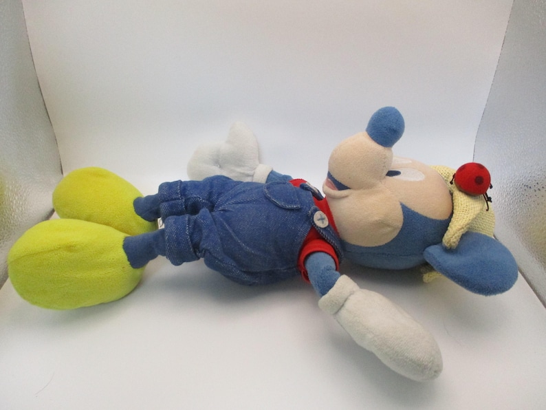May include: A plush toy of a cartoon character, lying on its back. The character has a blue head and hat, a red nose, and a yellow and red hat. It wears blue overalls, a red shirt, white gloves, and yellow shoes.