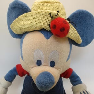May include: A plush toy mouse with blue ears, a beige face, and a yellow crocheted hat. A red ladybug with black spots is attached to the hat. The mouse wears a red and blue outfit.