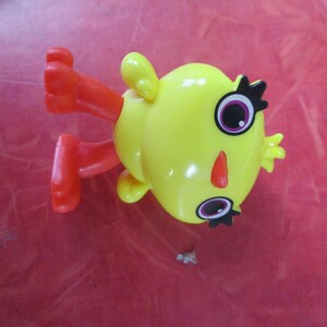 May include: A yellow toy figure with red legs and arms. The figure has large eyes with black eyelashes, a red nose, and small yellow ears. The toy is lying on a red surface.