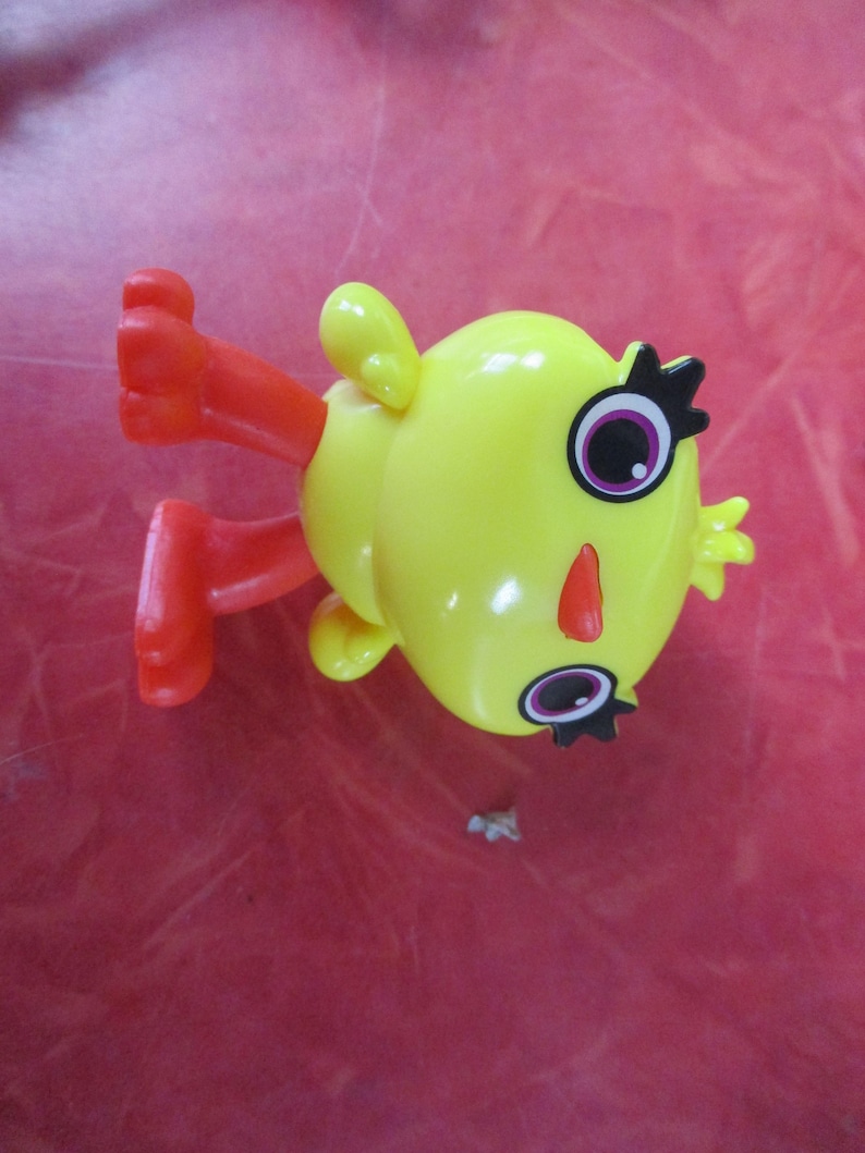 May include: A yellow toy figure with red legs and arms. The figure has large eyes with black eyelashes, a red nose, and small yellow ears. The toy is lying on a red surface.