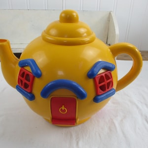 May include: A yellow plastic teapot with a lid, spout, and handle. The teapot is decorated with blue and red house-shaped details, including windows and a door. The teapot is sitting on a white surface.
