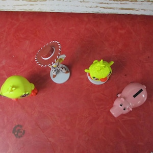 May include: A collection of small, colorful figurines on a red surface. The figurines include a pig-shaped bank, a character with a cowboy hat, and two green, spiky-haired figures. The toys are likely from a children's movie or TV show.