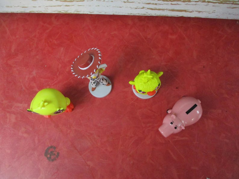 May include: A collection of small, colorful figurines on a red surface. The figurines include a pig-shaped bank, a character with a cowboy hat, and two green, spiky-haired figures. The toys are likely from a children's movie or TV show.