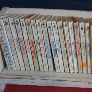 May include: A collection of vintage paperback books with white spines and titles in black and red text. The books are arranged on a white shelf, with titles including "Emma Darcy" and "Anne Mather".