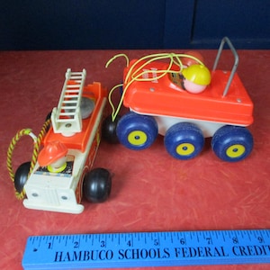 Könnte beinhalten: Zwei Oldtimer-Spielzeugfahrzeuge: ein Feuerwehrauto und ein Anhänger. Das Feuerwehrauto ist weiß und orange mit einer Leiter und einer Feuerwehrmannfigur. Der Anhänger ist orange mit blauen Rädern und einer Figur mit gelbem Hut. Beide Spielzeuge haben schwarze Räder.