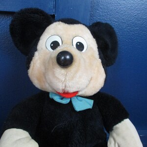 Op de afbeelding: Een pluche Mickey Mouse-speelgoed met een zwart lichaam, grote zwarte oren en een beige gezicht. Het speelgoed heeft een zwarte neus, witte ogen en een rode mond. Een lichtblauwe vlinderdas zit om de nek. Een klassiek Disney-personage.