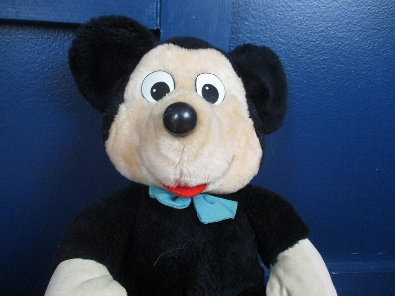 Op de afbeelding: Een pluche Mickey Mouse-speelgoed met een zwart lichaam, grote zwarte oren en een beige gezicht. Het speelgoed heeft een zwarte neus, witte ogen en een rode mond. Een lichtblauwe vlinderdas zit om de nek. Een klassiek Disney-personage.