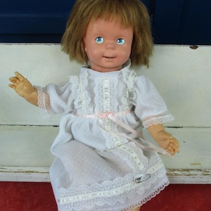 May include: A vintage doll with blonde hair and blue eyes, wearing a white dress with lace trim and a pink ribbon. The doll is seated and has a smiling expression.
