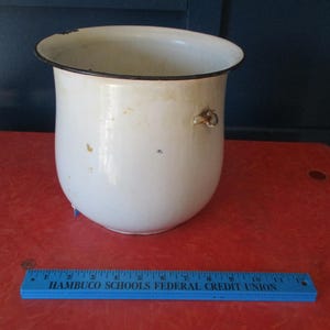 Könnte beinhalten: Ein weißer Emaille-Topf mit schwarzem Rand und kleinem Griff. Der Topf hat einen abgerundeten Körper und eine leicht ausgestellte Oberseite. Er steht auf einer roten Oberfläche. Ein blaues Lineal befindet sich im Vordergrund.