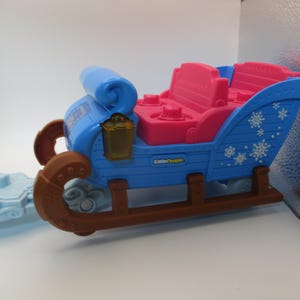May include: A blue and pink toy sleigh with brown runners and decorative snowflake accents. The sleigh has a blue seat back and a pink seat. A light blue sled is attached to the front.