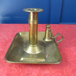 Puede incluir: Un candelero de latón antiguo con base cuadrada y copa cilíndrica alta. Una pequeña lámpara de aceite en forma de campana con asa se encuentra en la base. El metal tiene un cálido tono dorado.