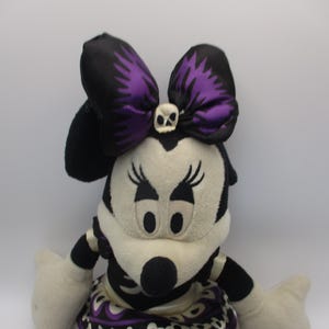 Puede incluir: Mu&ntilde;eca de peluche de Minnie Mouse en blanco y negro, con un gran lazo con detalles morados y un detalle de calavera. La mu&ntilde;eca tiene nariz negra, pesta&ntilde;as y una falda estampada.
