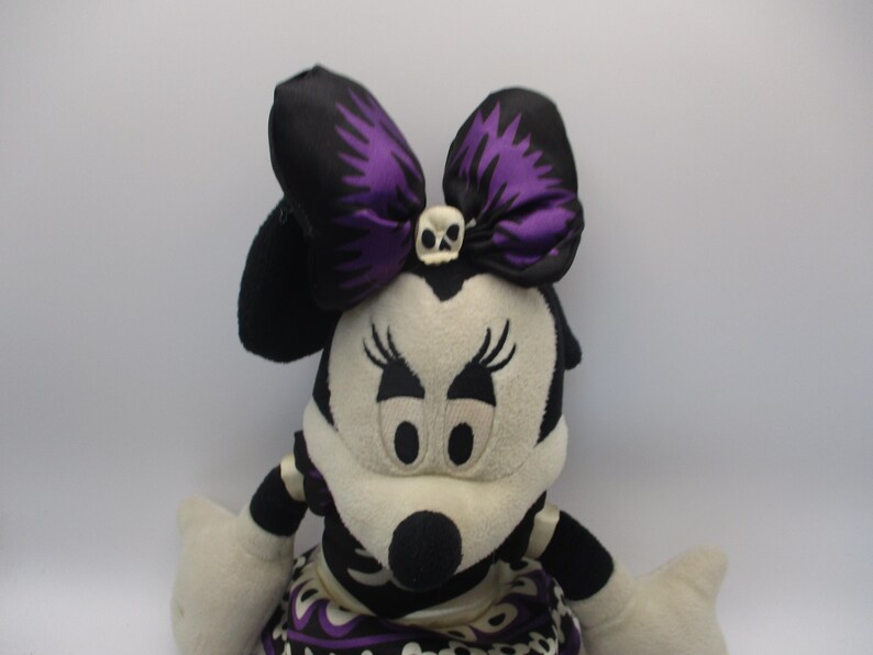 Puede incluir: Mu&ntilde;eca de peluche de Minnie Mouse en blanco y negro, con un gran lazo con detalles morados y un detalle de calavera. La mu&ntilde;eca tiene nariz negra, pesta&ntilde;as y una falda estampada.