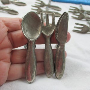 May include: A set of vintage silver-toned miniature cutlery, including a spoon, fork, and knife. The spoon, fork, and knife are held together. Additional cutlery pieces are visible in the background. The items are on a white surface.