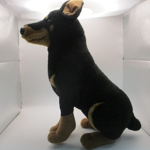 May include: A plush black and tan Doberman Pinscher dog toy. The stuffed animal has tan markings on its snout, ears, chest, and paws. The dog is sitting upright with its tail curled.