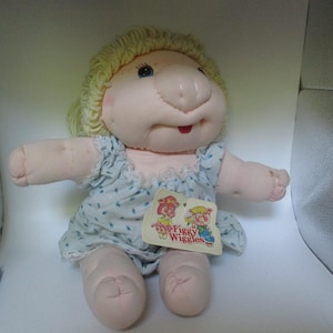 Op de afbeelding: Een zachte stoffen pop met blond garen haar, een ronde neus en een lichtroze huidskleur. De pop draagt een lichtblauwe mouwloze romper met een klein blauw blaadjespatroon en witte kanten rand. Een label aan de romper bevat de tekst Figgy Wiggles.