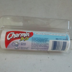 Könnte beinhalten: Eine zylindrische Rolle Charmin To Go Toilettenpapier in einer transparenten Kunststoffverpackung. Das Etikett enthält roten und blauen Text, einen Strichcode sowie Anleitungen mit Illustrationen zum Öffnen der Kunststofflaschen.