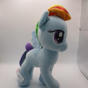 May include: A light blue plush pony with a rainbow-colored mane and a yellow and red hat. The pony has purple eyes, a purple tail, and sparkly blue wings. A white tag is attached to the pony's foot.