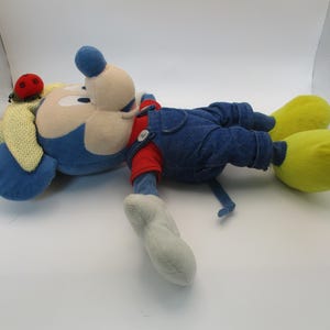 May include: A plush toy of a cartoon mouse, lying on its side. The mouse has a blue head and ears, a beige face, and is wearing blue denim overalls over a red shirt. Yellow feet and a ladybug on the ear complete the look.