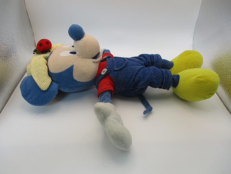 May include: A plush toy of a cartoon mouse, lying on its side. The mouse has a blue head and ears, a beige face, and is wearing blue denim overalls over a red shirt. Yellow feet and a ladybug on the ear complete the look.