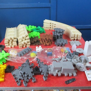 May include: A collection of colorful plastic building blocks and track pieces, including beige ramps, green and brown textured blocks, and gray interlocking components. Various other pieces, such as wheels and figures, are also present, suggesting a construction toy set.