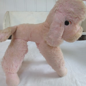 May include: A vintage pink plush poodle toy with black and red eye details. The poodle has a fluffy head, long ears, and a tail. The toy is made of a soft, fuzzy material and is standing on its four legs.