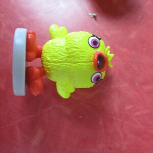 May include: A yellow chick figurine with red feet and a red beak. The chick has large pink and black eyes and a textured body. A gray base supports the figurine.