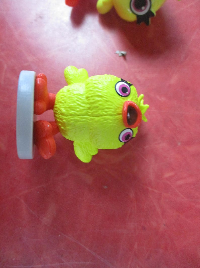 May include: A yellow chick figurine with red feet and a red beak. The chick has large pink and black eyes and a textured body. A gray base supports the figurine.