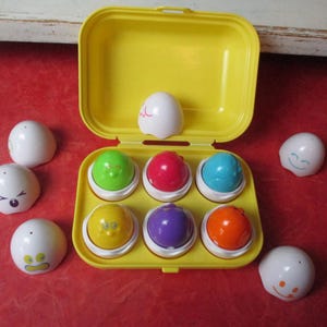 May include: A yellow plastic egg-shaped carrying case, open to reveal six colorful eggs. Each egg has a different color and a smiling face. Several white eggs with different facial expressions are scattered around the case.