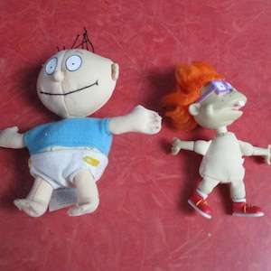 May include: Two plush dolls of cartoon characters. One doll has a cream-colored face, blue shirt, and white diaper. The other doll has orange hair, purple glasses, and a white outfit with red shoes. The dolls are on a red surface.