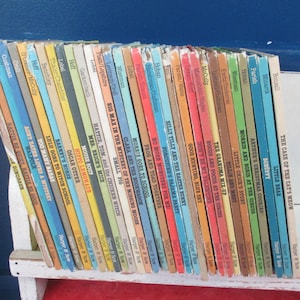 Puede incluir: Una colección de libros infantiles antiguos con lomos coloridos, dispuestos sobre una estantería blanca sobre un fondo azul. Los títulos son visibles, mostrando una variedad de historias. Los libros están en buen estado, lo que sugiere que se han conservado bien.