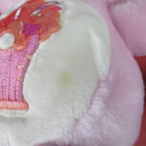 May include: Close-up of a pink plush toy with a white patch featuring an embroidered design. The design includes a pink and orange sundae with a heart and a white circle. The plush has a soft, fuzzy texture.