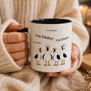 Puede incluir: Una taza de cerámica blanca con interior y asa negros, que presenta un gráfico de cuatro gaviotas blancas y negras con los picos abiertos. El texto "Frau Schubert" está impreso en negro en varios tamaños sobre las ilustraciones de las aves.