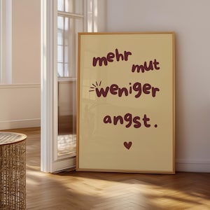 Op de afbeelding: Een ingelijste poster met de woorden "mehr mut weniger angst" in een donkerrood lettertype, samen met een klein hartje. De poster staat in een kamer met een houten vloer en een rotan tafel.