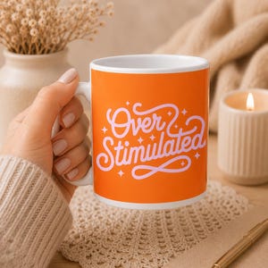 Könnte beinhalten: Eine weiße Keramiktasse mit orangefarbenem Hintergrund und dem weißen Schriftzug "Over Stimulated". Die Tasse hat einen weißen Henkel. Die Tasse steht auf einer Holzoberfläche.