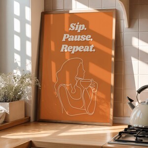 May include: Framed orange art print with the words "Sip. Pause. Repeat." in white. A white line drawing of a person holding a cup is below the text. The print is displayed on a wooden countertop.
